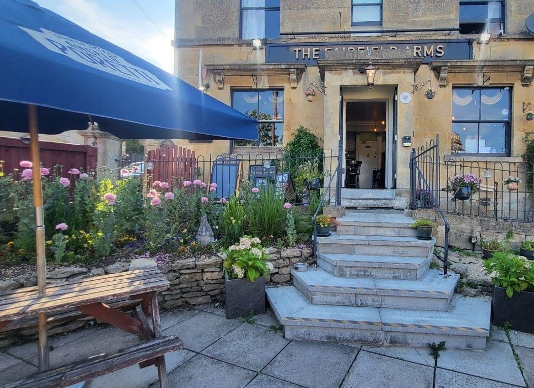 The Bath stone front of the pub in summer. The name is in gold capitals and stone steps lead up to the open door past a bed full of flowers.