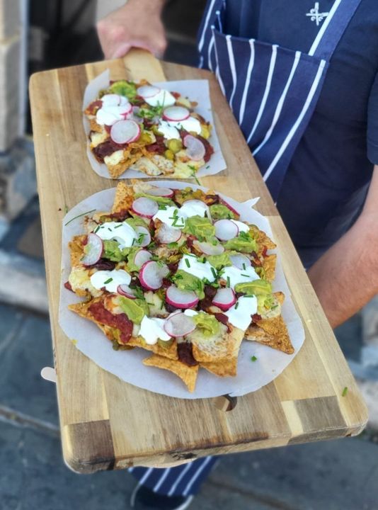 Two plates of nachos on a long wooden board, carried out by someone in a striped apron. They are topped with melted cheese, guacamole, sour cream, sliced radish and chives.