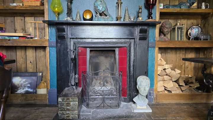 The fireplace by daylight: a black iron surround with red tiles and a fireguard, and logs stacked on the shelves beside it. Busts and curiosities stand on the mantel against the teal wall, and another bust sits on the hearth.