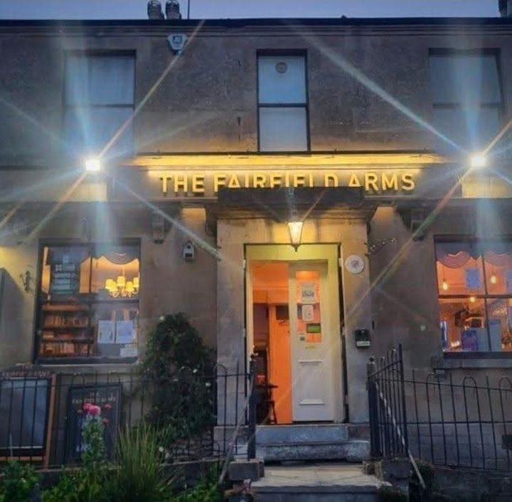 The front of the pub at dusk with its lamps lit. The name glows in gold capitals above the open door, and the windows on either side are lit from within.